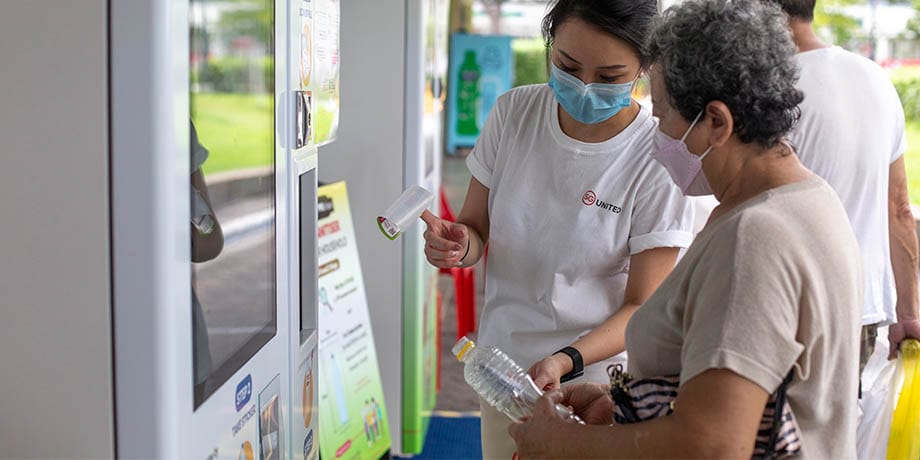 Volunteer helping old lady with hand sanitiser collection from smart vending machine 