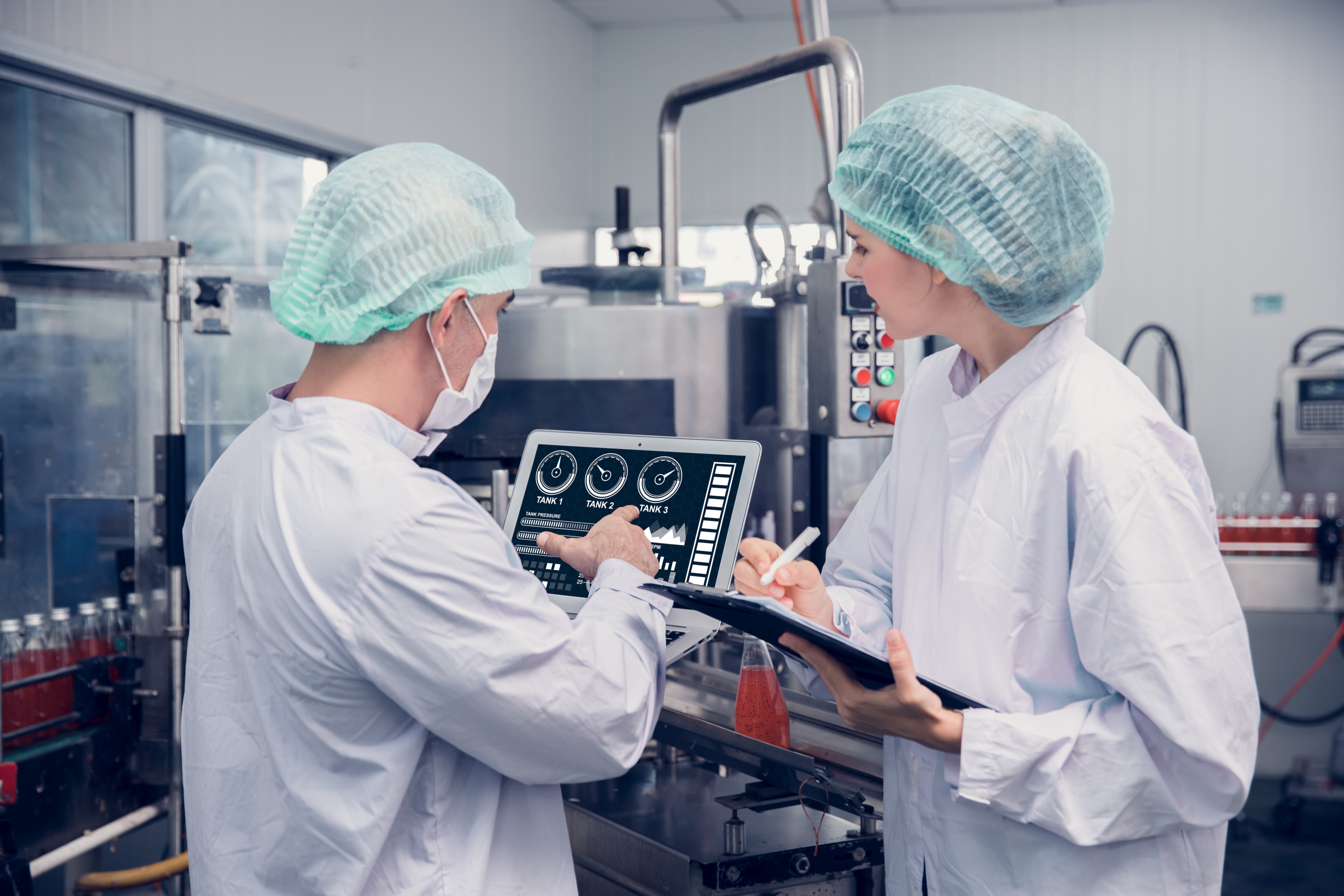 Two industrial workers, a man and a woman, wearing hard hats and safety vests, looking at a tablet.